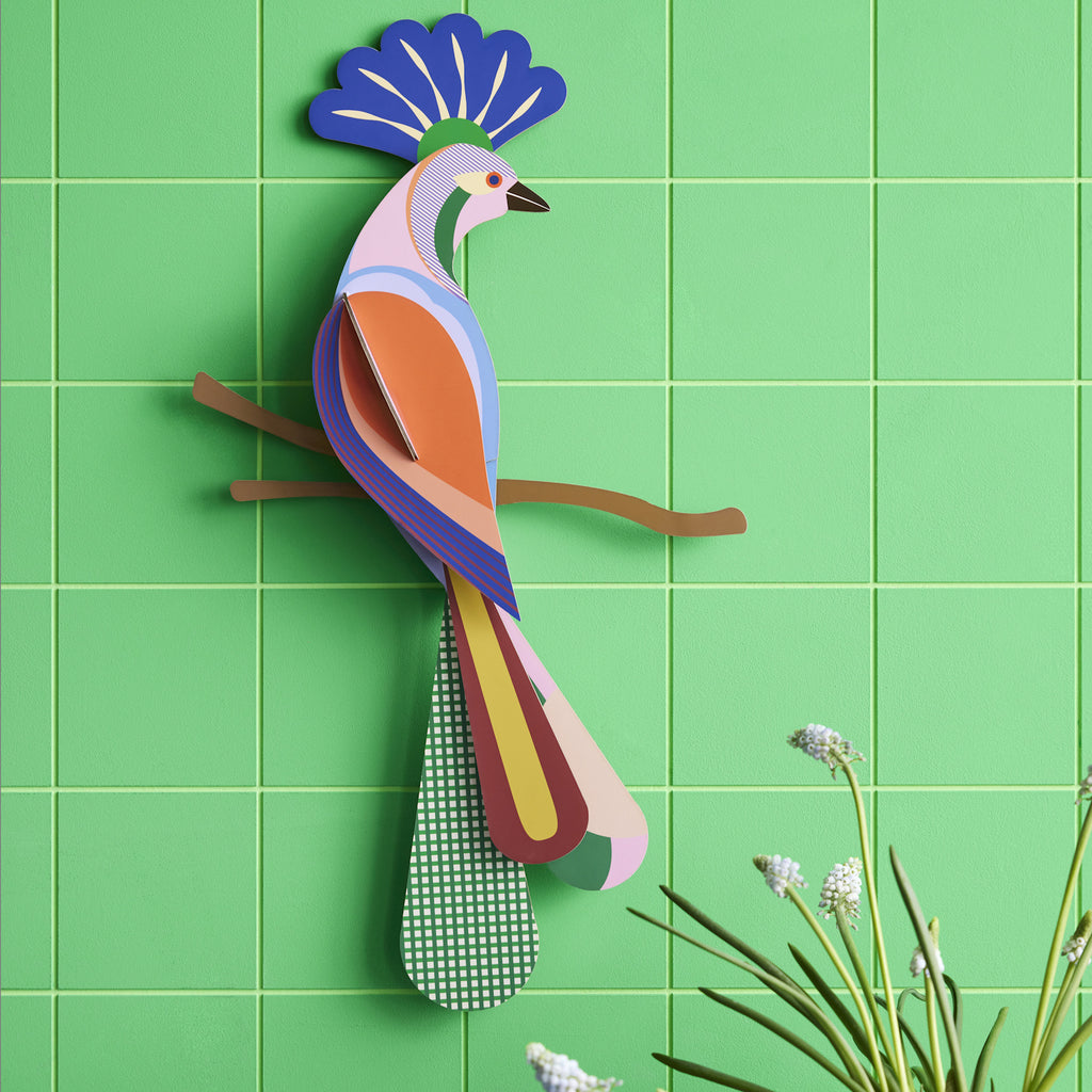 A colorful, stylized bird art sculpture on a light green tiled wall. The bird is perched on a branch, with a vibrant plumage and geometric patterns.