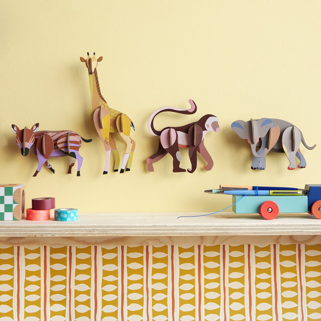 Four colorful, paper-cut animal figures, a gazelle, giraffe, monkey, and elephant, are mounted on a light yellow wall. A playful, artistic tone.