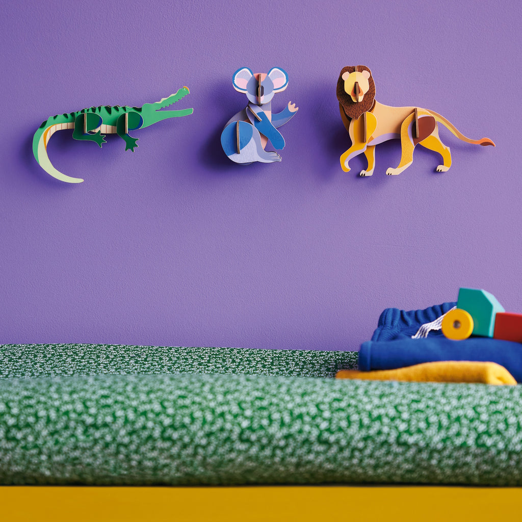 Colorful wall with crocodile, koala, and lion 3D art on a purple background. Below, a textured green surface with folded clothes and toy blocks. Playful and vibrant.