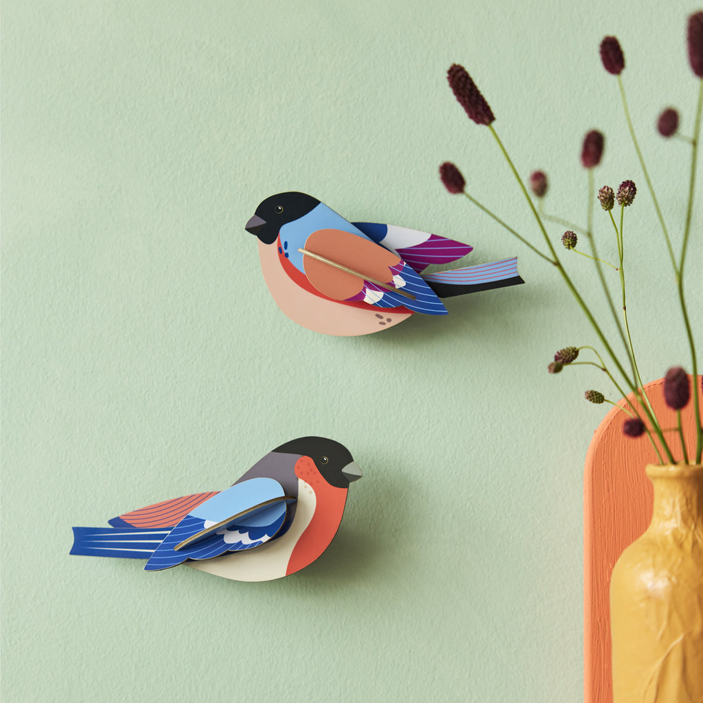 Paper craft birds with vibrant colors, including blue, orange, and pink, adorn a light green wall. A yellow vase with delicate purple flowers adds a warm touch.