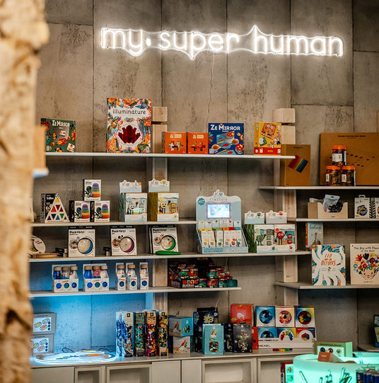 Shop shelves beneath a glowing "my super human" neon sign, displaying colourful children's toys and games including Illuminature and Ze Mirror. 