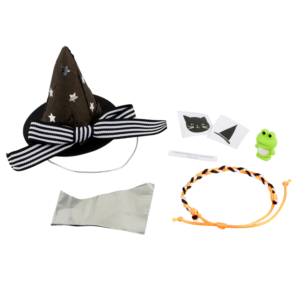 A black witch hat with white stars and a striped bow, a frog figurine, cat and hat stickers, a braided orange cord, and a silver fabric are laid out on a white background.