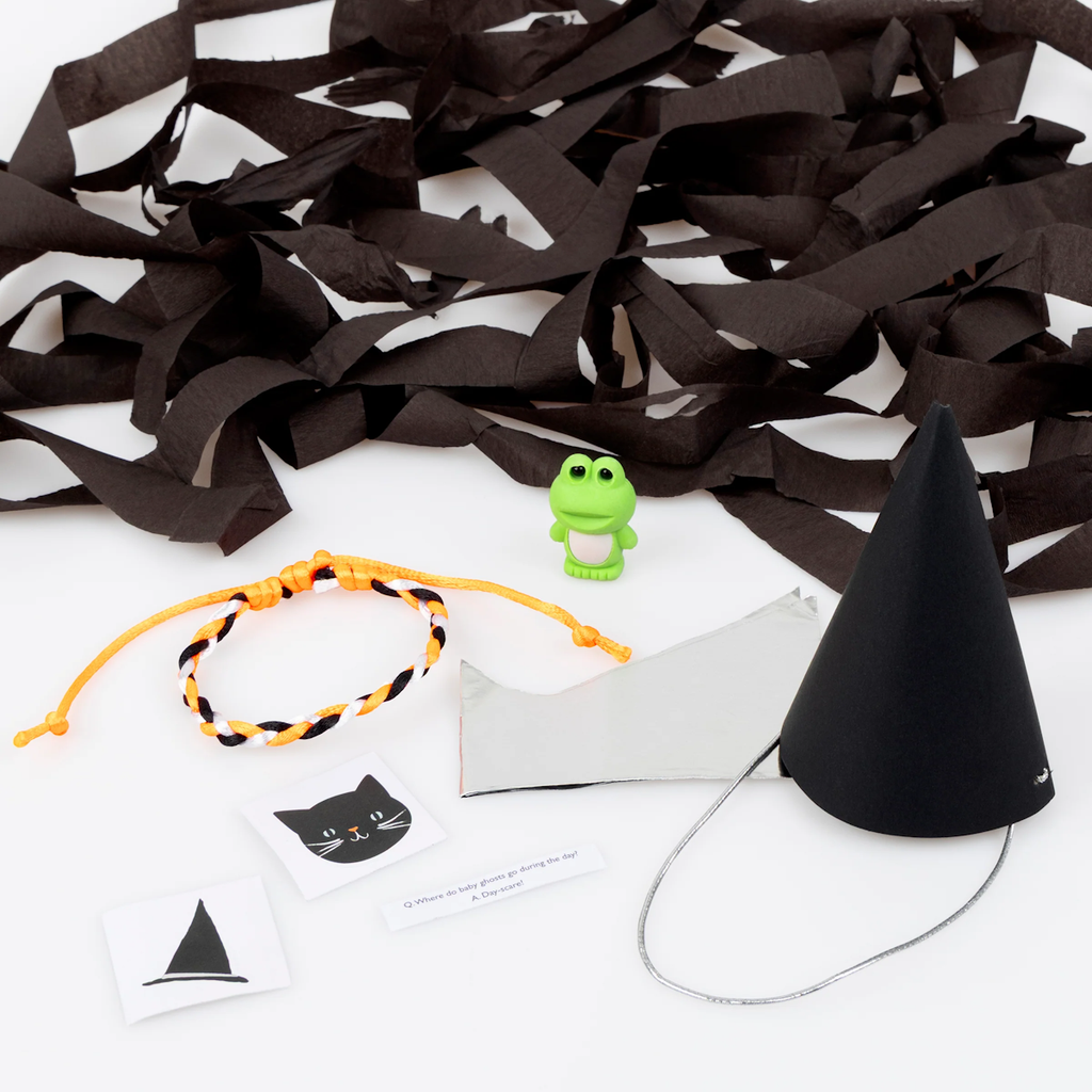 Assorted Halloween-themed items include black streamers, a green frog toy, a black cone hat, orange braided bracelet, and cat and witch hat cards.