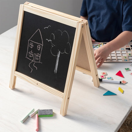 Child playing at a table with a wooden blackboard easel featuring a chalk drawing of a house and tree.