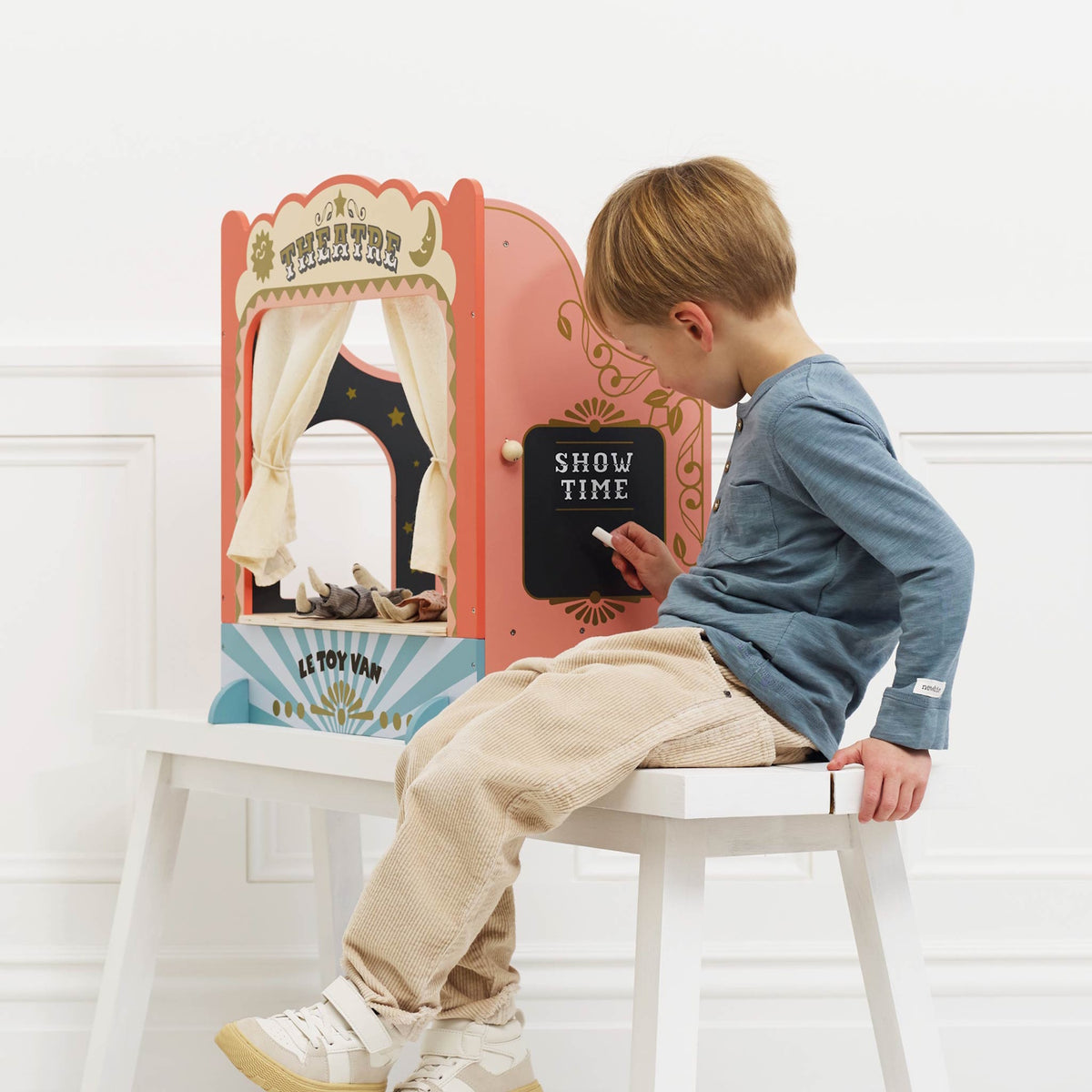 Child sitting on a bench writing on the side chalkboard of a tabletop wooden puppet theatre.