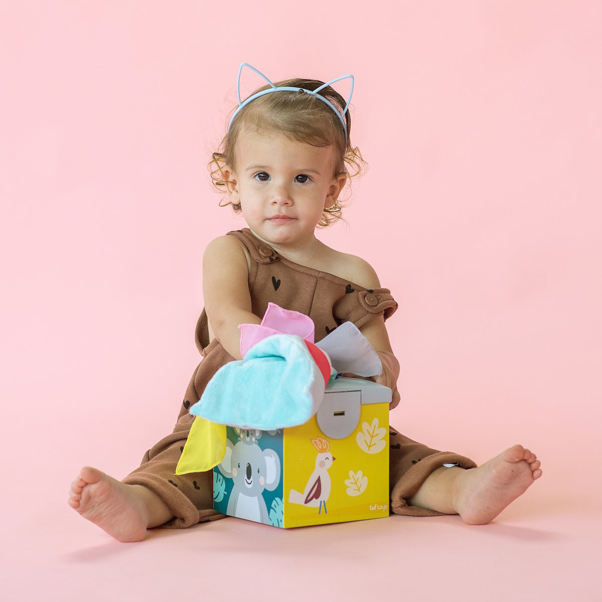 Toddler wearing cat ears headband sitting on pink background, pulling fabrics from Kimmy Koala Wonder Tissue Box.