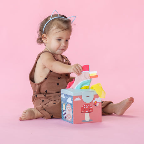 Toddler wearing cat ears sitting on a pink background, pulling a sensory cloth from a colorful toy box.
