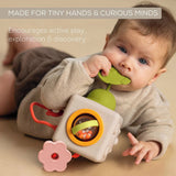 Baby lying on tummy chewing a green teether attached to a soft sensory activity cube toy.
