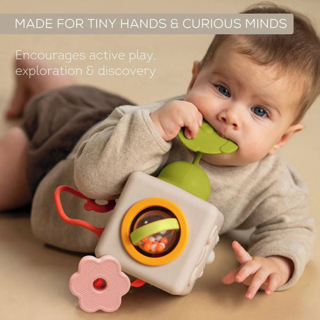 Baby lying on tummy chewing a green teether attached to a soft sensory activity cube toy.