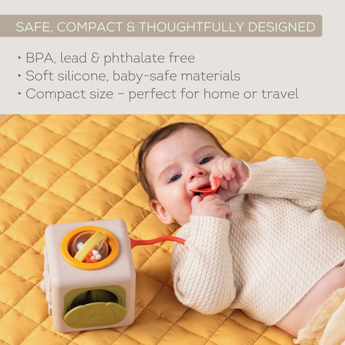 Baby lying on a yellow mat chewing a soft silicone toy attached to an activity cube.