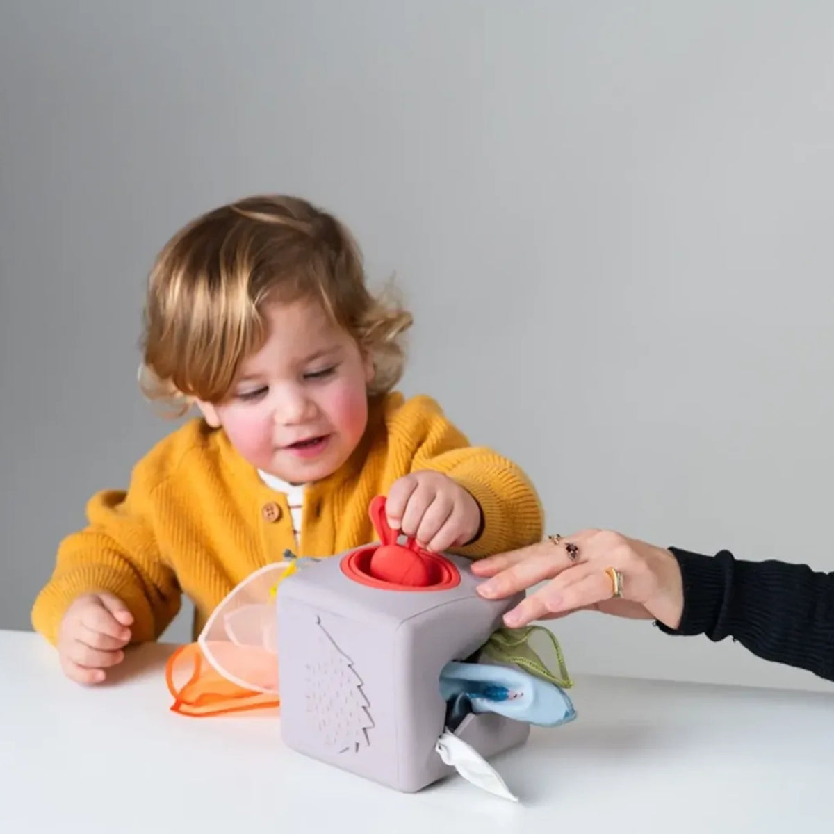 Toddler in yellow cardigan playing with a Taf Toys pop-up tissue box toy at a table.