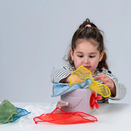 Toddler playing with a sensory tissue box toy, pulling out colorful fabric tissues at a table.