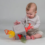 A baby sitting on the floor playing with a gray pop-up sensory tissue box toy with colorful sheer fabrics.