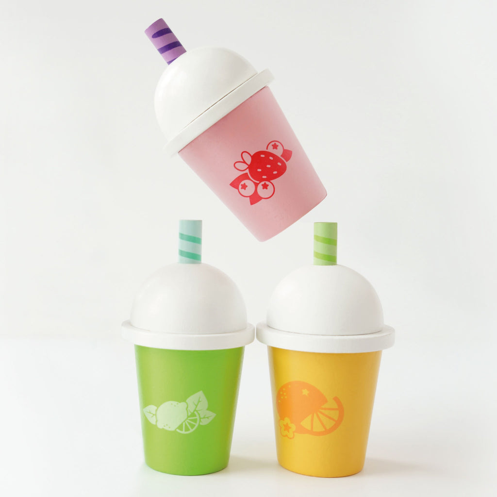 Three colorful cups with white domed lids and striped straws. Each cup has fruit designs: orange on yellow, strawberry on pink, lime on green. Vibrant and playful.
