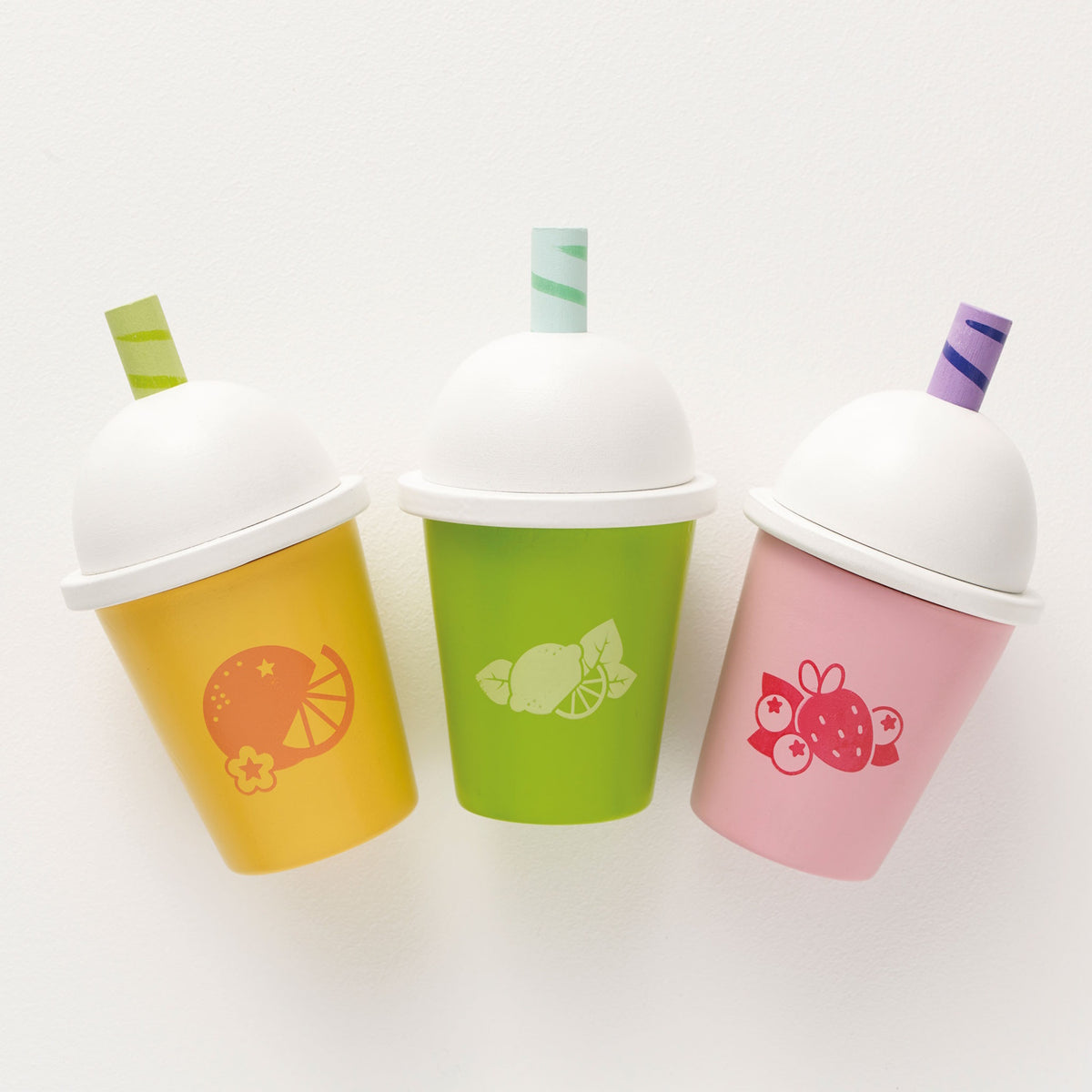 Overhead view of three wooden toy smoothie cups in yellow, green, and pink on a white surface.