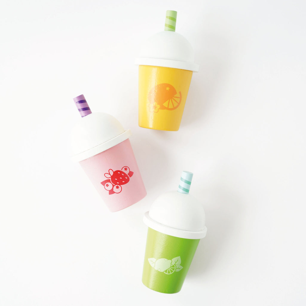 Three colorful cups with white domed lids and striped straws. Each cup has fruit designs: orange on yellow, strawberry on pink, lime on green. Vibrant and playful.