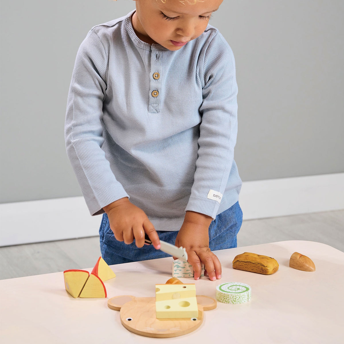 Child wearing a grey button-down shirt playing with a wooden cheese chopping board set and a toy knife.