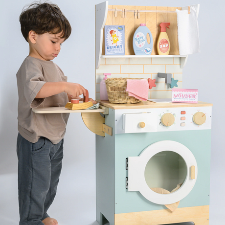 Young boy using a wooden toy iron on the fold-out side board of a play laundrette set.