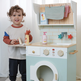 Toddler boy holding wooden spray bottle and iron next to a play laundrette set.