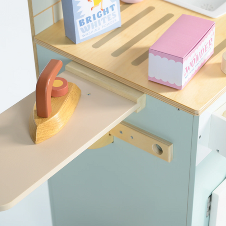 Close-up of a wooden toy iron on a foldable ironing board with toy detergent boxes.