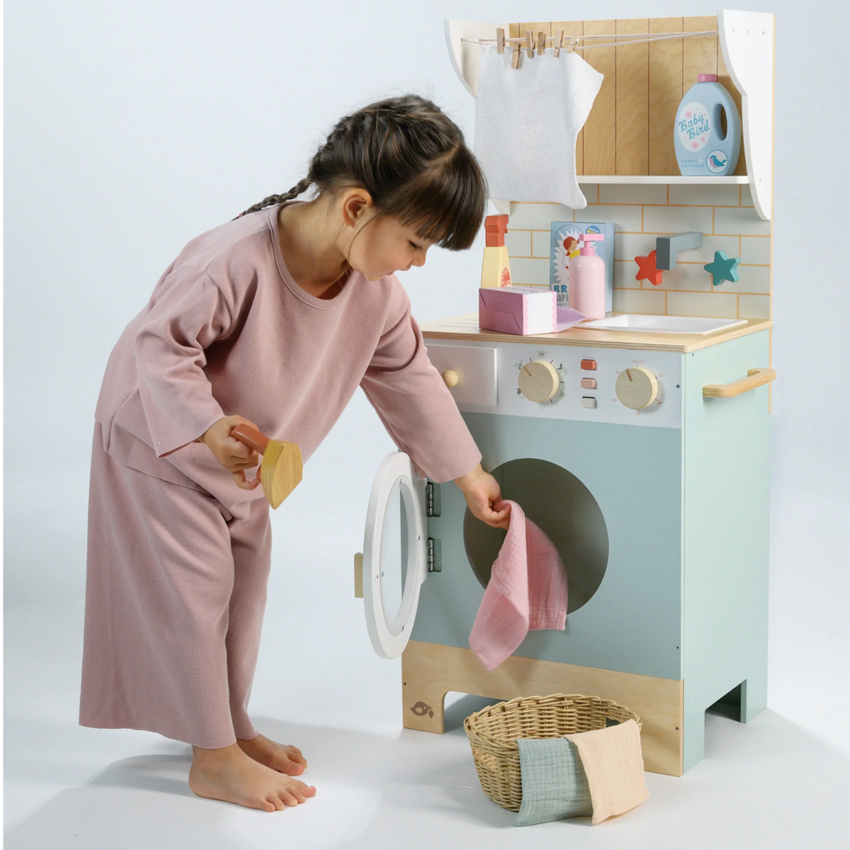 Little girl putting play laundry into the wooden washing machine while holding a toy iron.