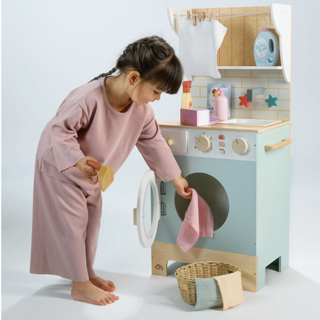 Little girl putting play laundry into the wooden washing machine while holding a toy iron.
