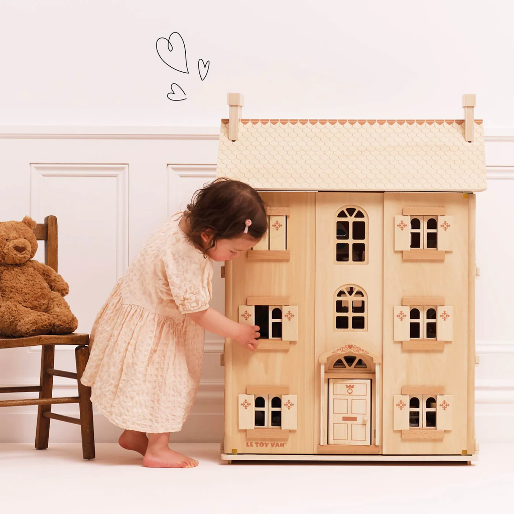 A young child in a cream dress plays with a wooden dollhouse. A teddy bear sits on a chair nearby. The scene is warm and playful, with drawn hearts above.