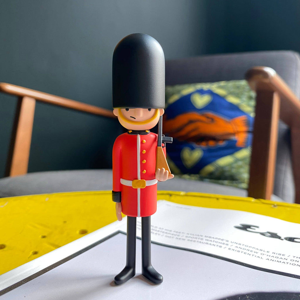 Toy figure of a British guard in a red uniform and tall black hat stands on a table. Behind is a couch with a cushion featuring a handshake design.