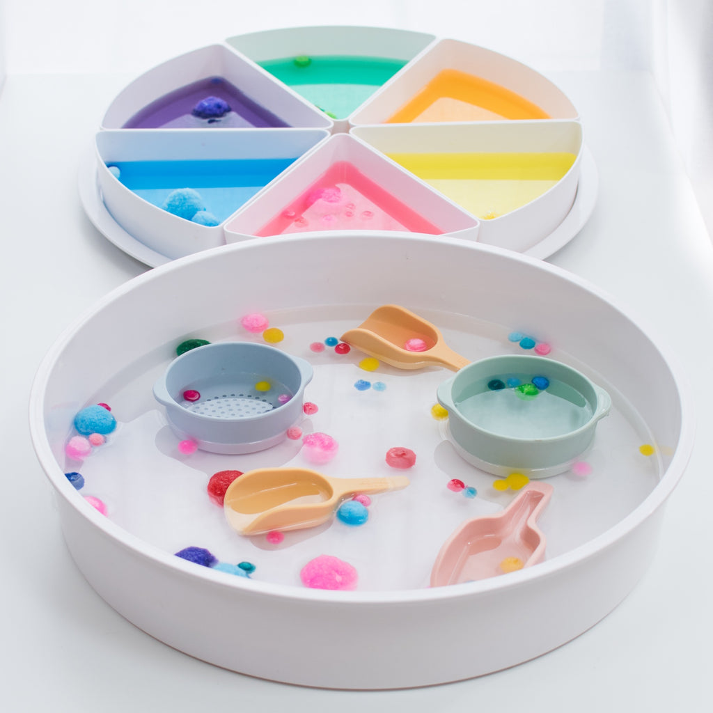 A white sensory play tray filled with colorful pom-poms, scoops, and cups. A nearby divided dish holds sections of textured materials in pastel hues.