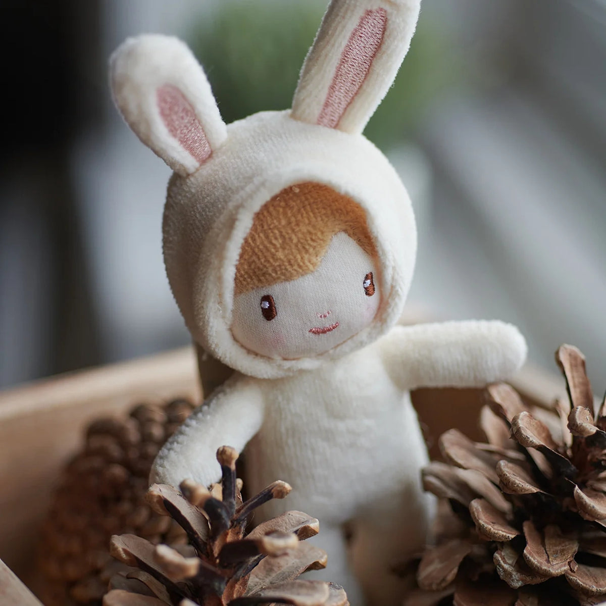 A close-up of a tiny plush doll wearing a cream bunny costume surrounded by pine cones.