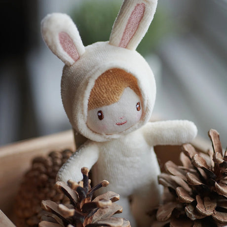 A close-up of a tiny plush doll wearing a cream bunny costume surrounded by pine cones.