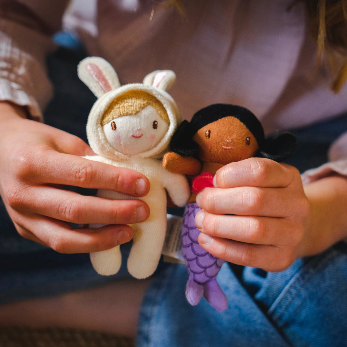 Child hands holding a small plush doll in a bunny suit alongside a tiny purple mermaid doll.