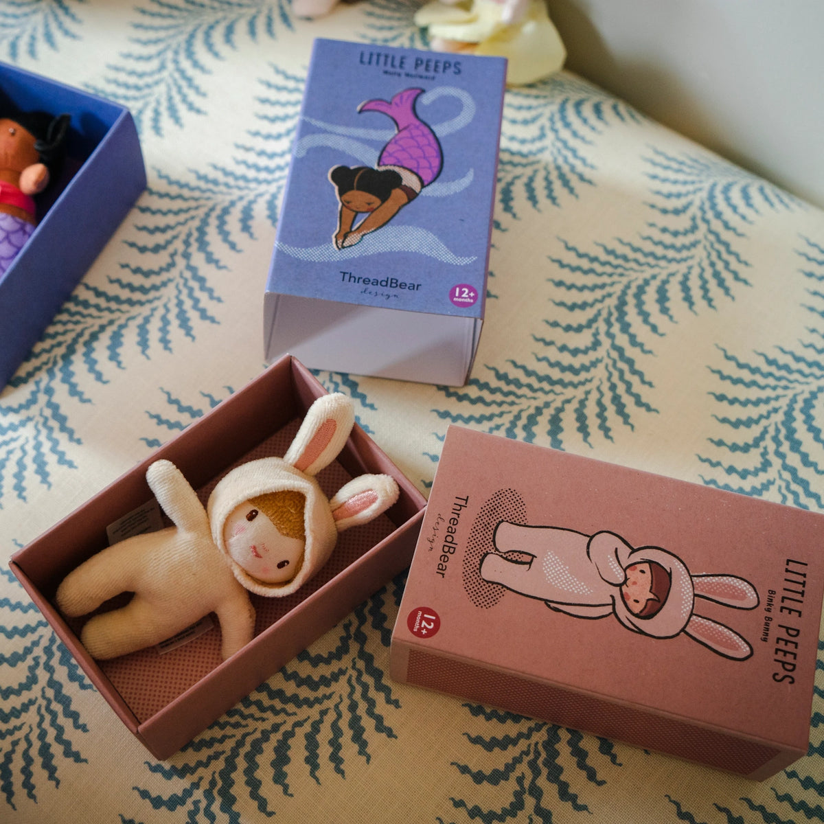 Small plush doll in a bunny outfit resting inside an open pink Little Peeps box on a pattern fabric.