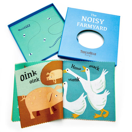 Open fabric book titled "The Noisy Farmyard" featuring illustrations of pigs and geese alongside its blue gift box.