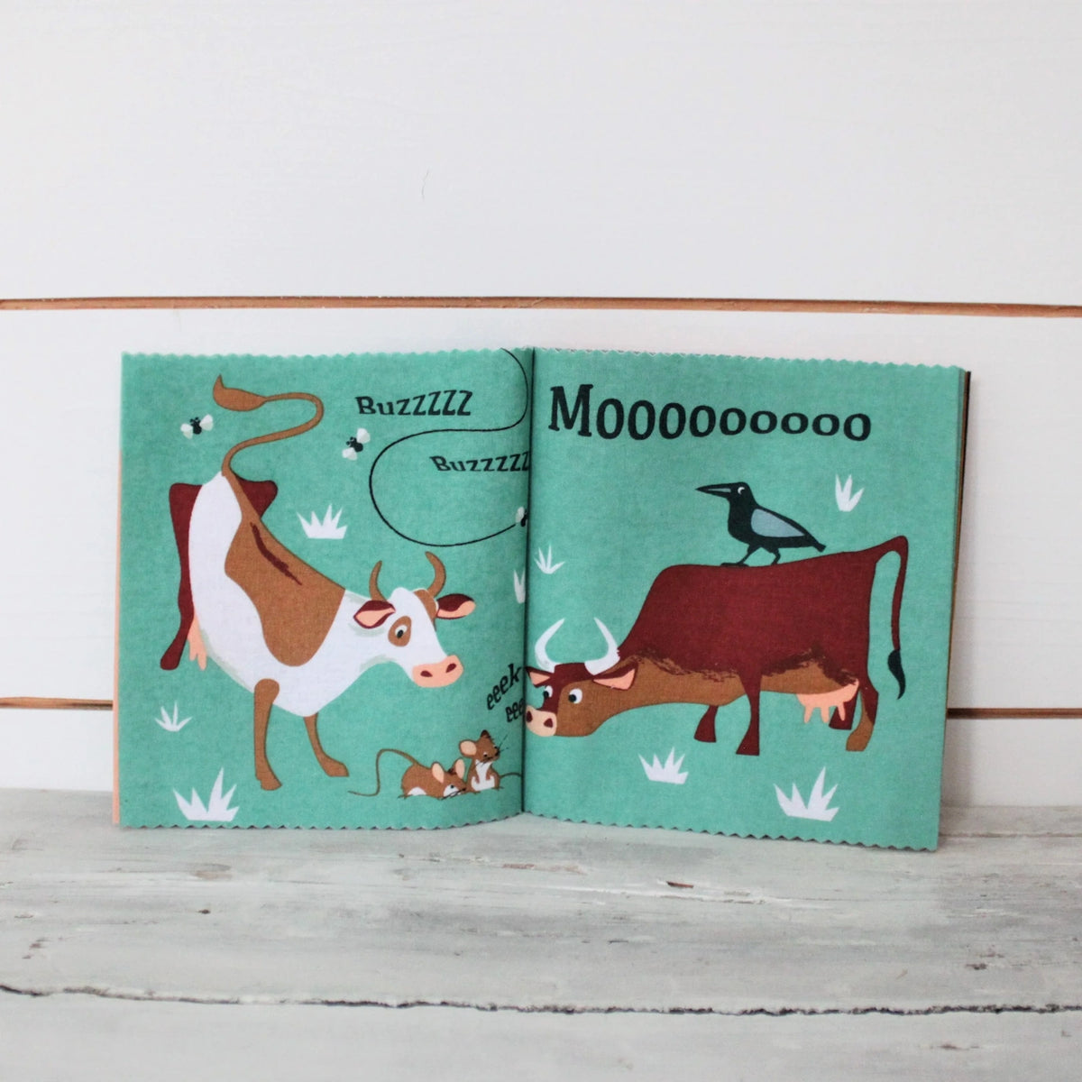 Open fabric book showing cows, mice, flies, and a bird on teal pages.