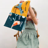Child in green overalls holding up an open fabric book featuring a donkey and an owl.