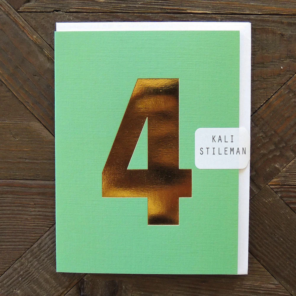 A green card featuring a large, shiny gold number "4" in the center. A small white label on the right reads "Kali Stileman." Wood background.
