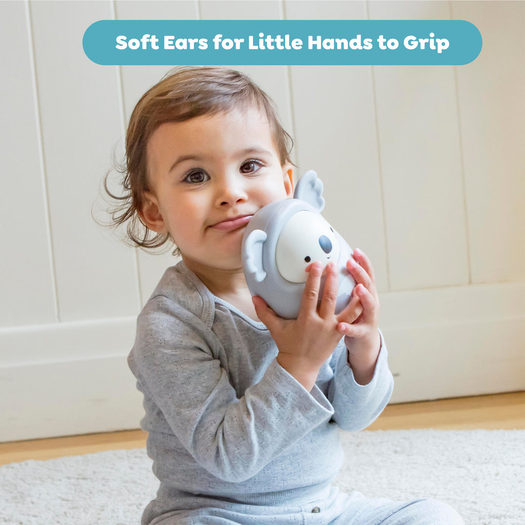 A toddler in grey pajamas holds a toy with soft ears, grinning slightly. The background is a white panel wall, conveying a cozy, playful mood. Text reads "Soft Ears for Little Hands to Grip."