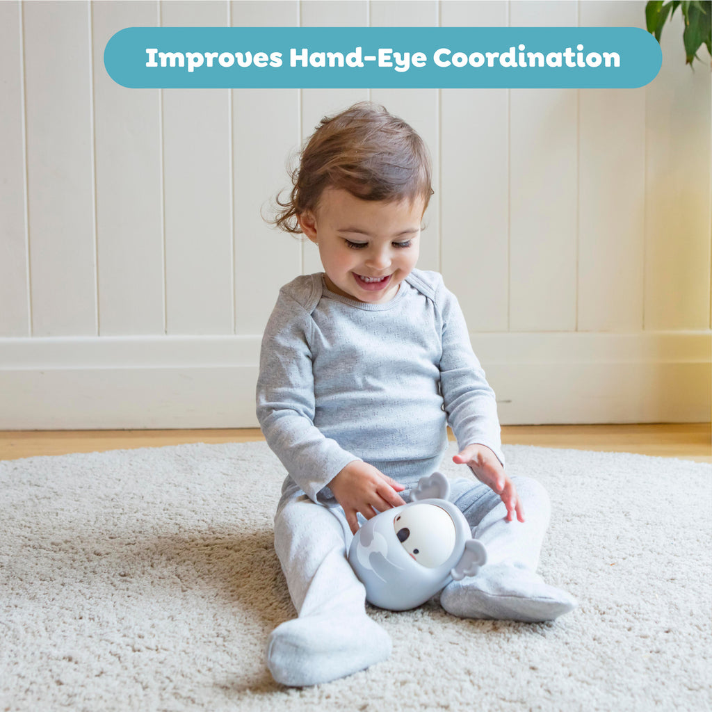 A toddler in gray pajamas sits on a beige carpet playing with a smiling koala toy. The mood is joyful and relaxed, highlighting hand-eye coordination.