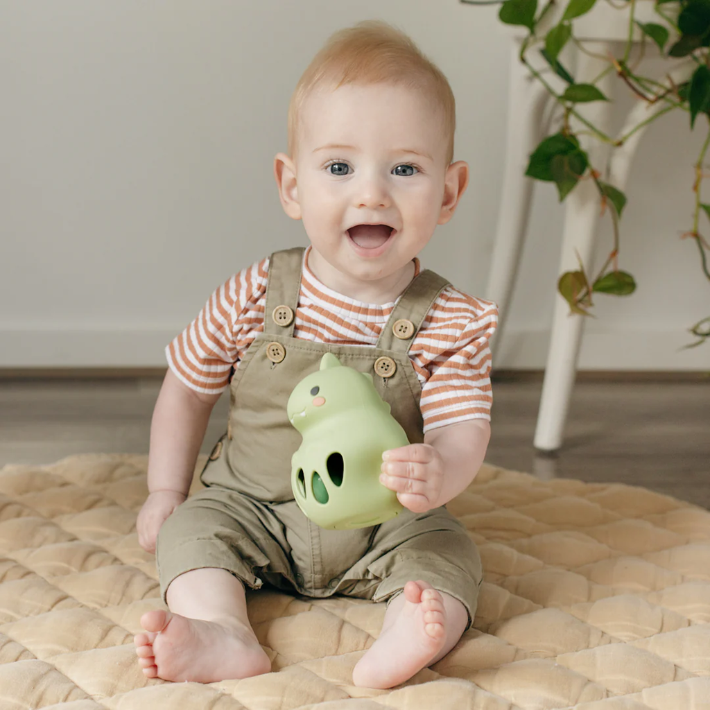 A happy baby with light brown hair, wearing a striped shirt and beige overalls, sits on a quilted mat holding a green toy. A plant is in the background.