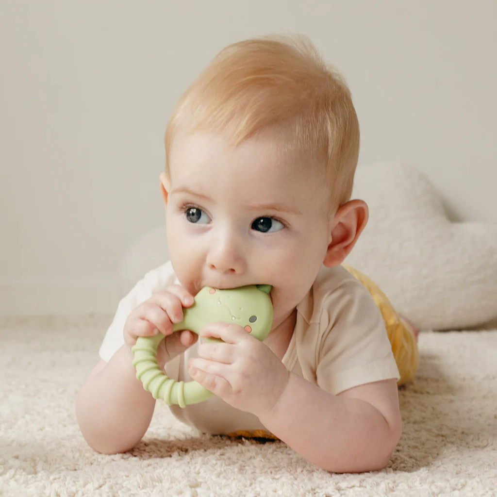 A baby with light hair is lying on a soft carpet, chewing on a green teething toy. The scene is calm and gentle, with a neutral background and soft plush decor.