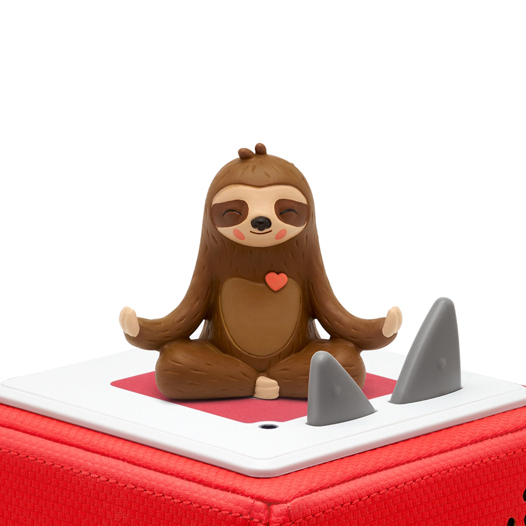 A cute sloth figurine with a serene expression sits cross-legged in a meditative pose, a small heart on its chest, atop a red toy box with gray controls.