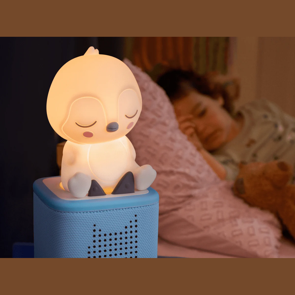 A glowing penguin-shaped night light sits on a blue speaker beside a sleeping child. The scene conveys warmth and tranquility, with a soft pink pillow and cozy ambiance.