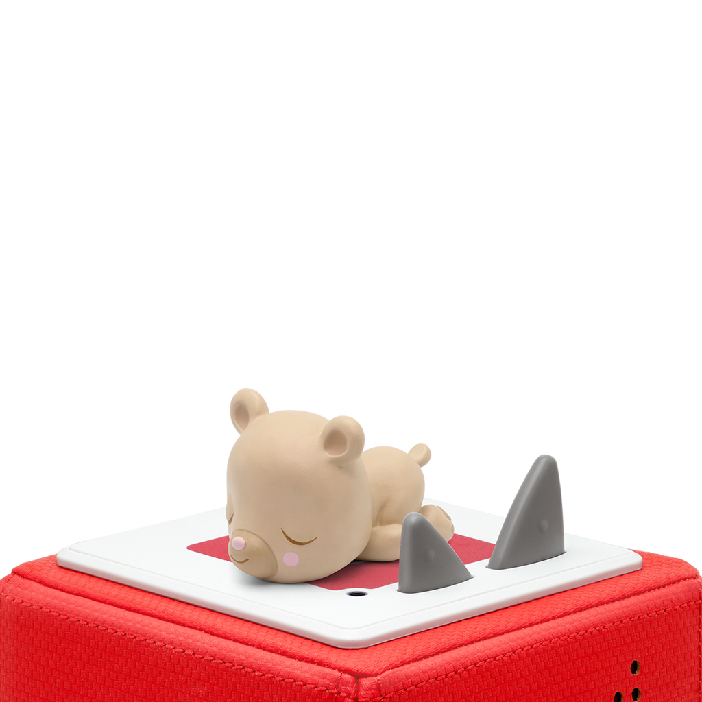 A cute, beige bear figurine peacefully sleeps atop a red speaker with grey triangular buttons, set against a white background. Cozy, serene mood.