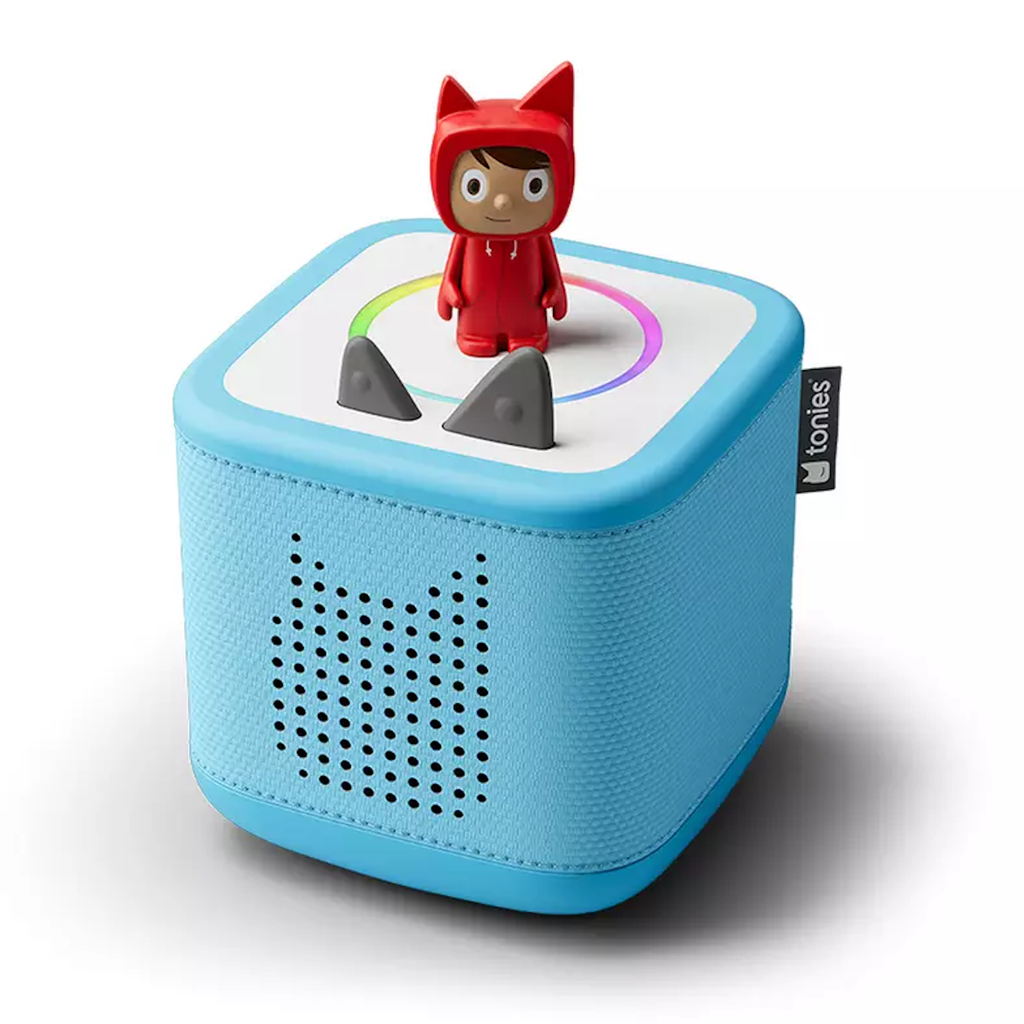 A blue Toniebox speaker with a cute character figurine in a red outfit stands on top. The box features two gray triangular buttons and a small speaker panel.