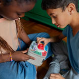 Two kids sitting on the floor using a red controller on a light gray Toniebox audio player together.