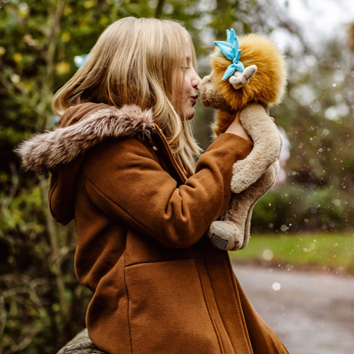 A young blonde girl in a winter coat holds up a plush creature, kissing its nose outdoors in a park.