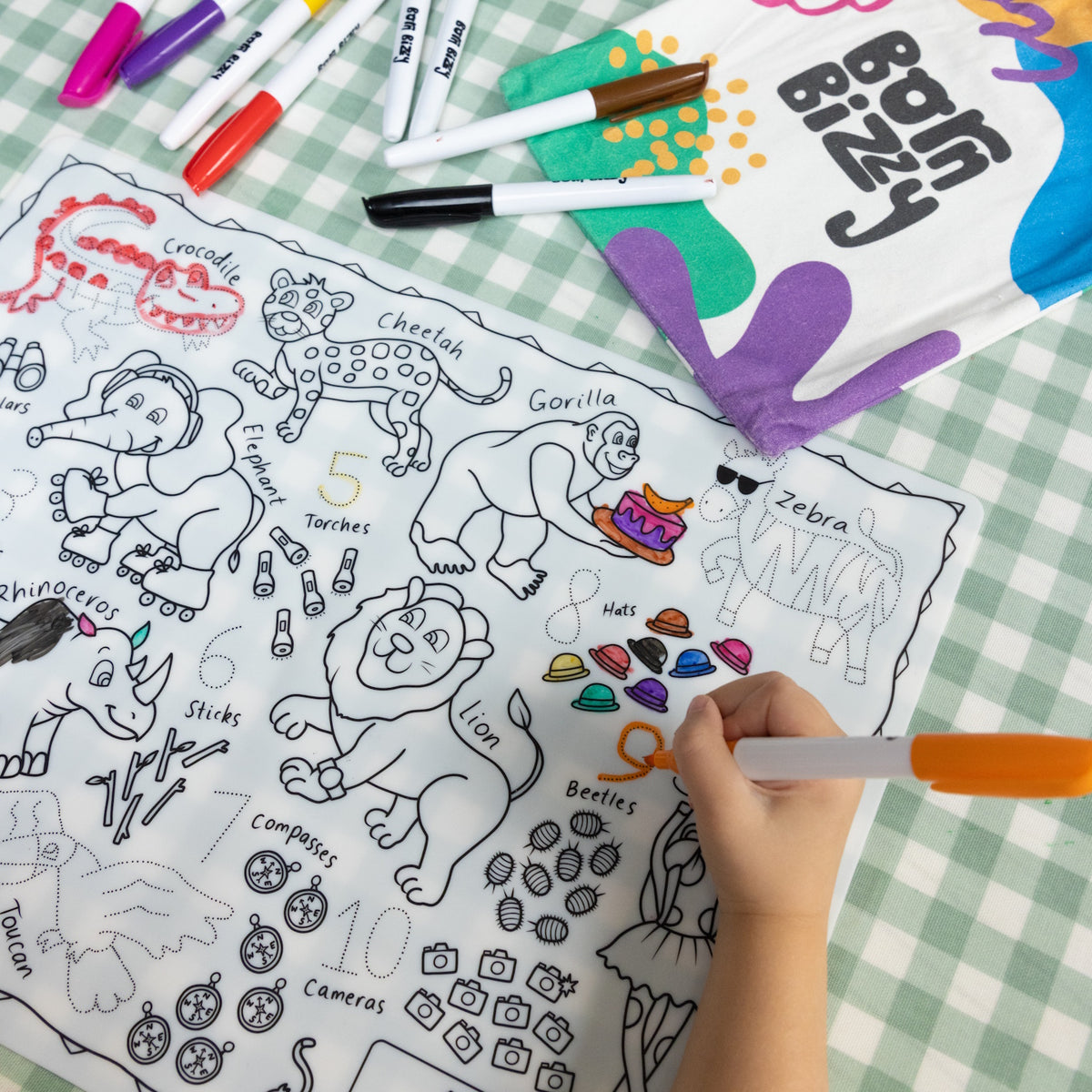 A child coloring an animal-themed activity mat with markers next to a "Born Bizzy" canvas bag.