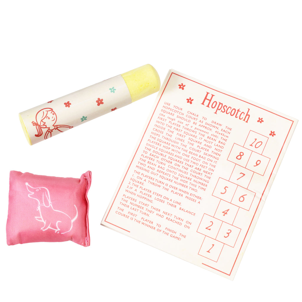 Hopscotch set with large yellow chalk, pink beanbag with dog illustration, and hopscotch rules card featuring numbered squares. Playful and nostalgic.