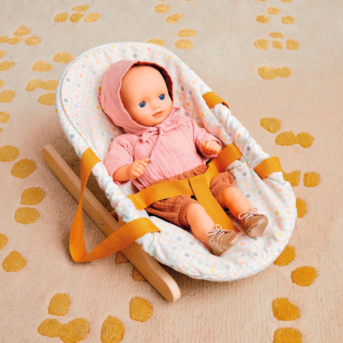 Baby doll in a pink hoodie sitting in a wooden toy bouncer with mustard yellow straps on a patterned rug.